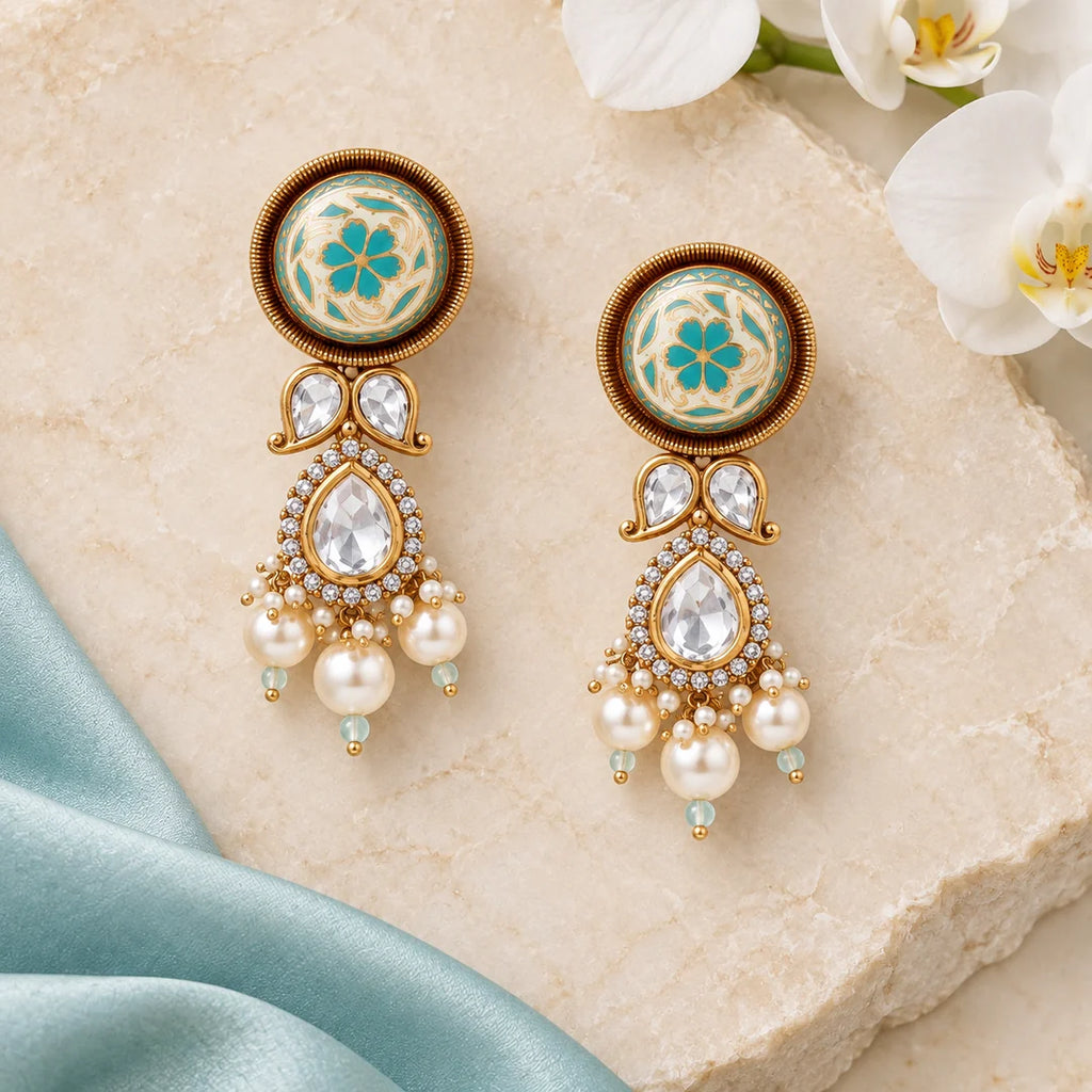 Pair of Malvika turquoise and ivory floral drop earrings arranged on cream stone beside pale aqua silk and white orchids.