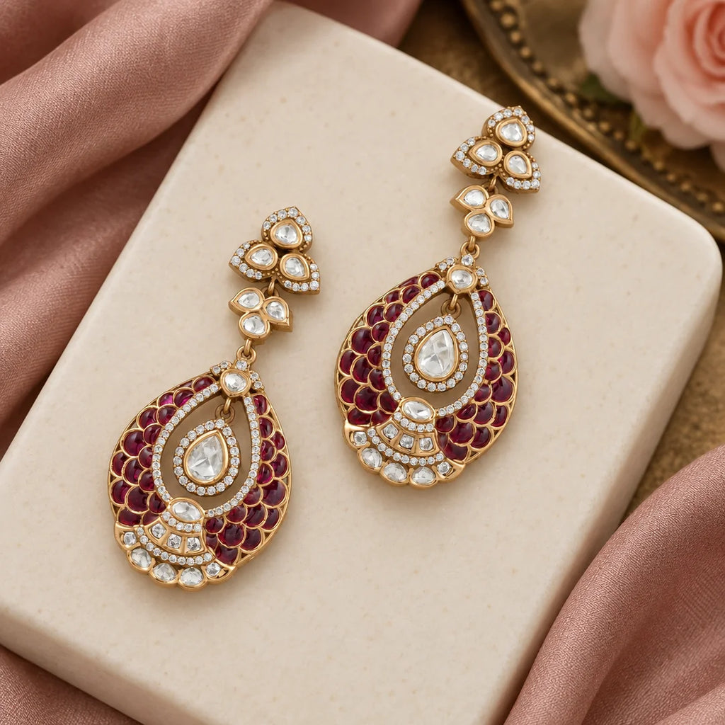 Pair of Mayura wine teardrop earrings on an ivory display surrounded by blush silk and a pale rose.