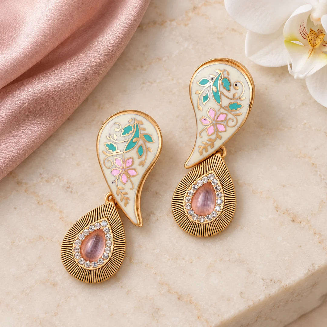 Pair of Pallavi ivory and pastel paisley earrings on cream stone beside blush silk and a white orchid.