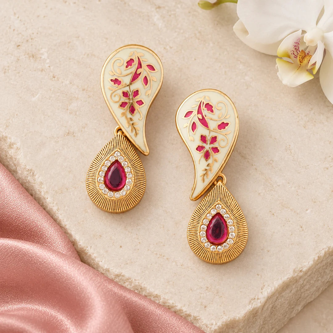 Pair of ivory and ruby-pink Pallavi paisley earrings on cream stone beside blush silk and a white orchid.