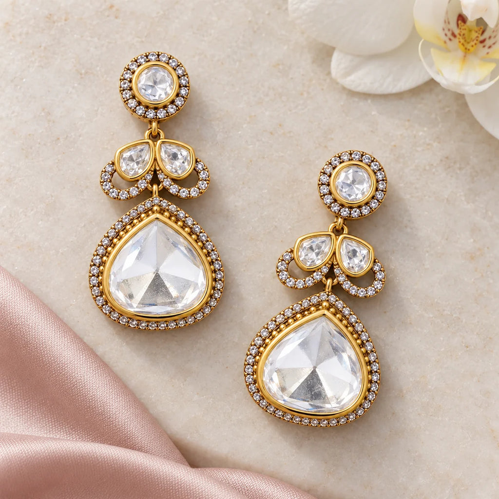 Pair of Prisha clear stone teardrop earrings on ivory stone beside blush silk and a white orchid.