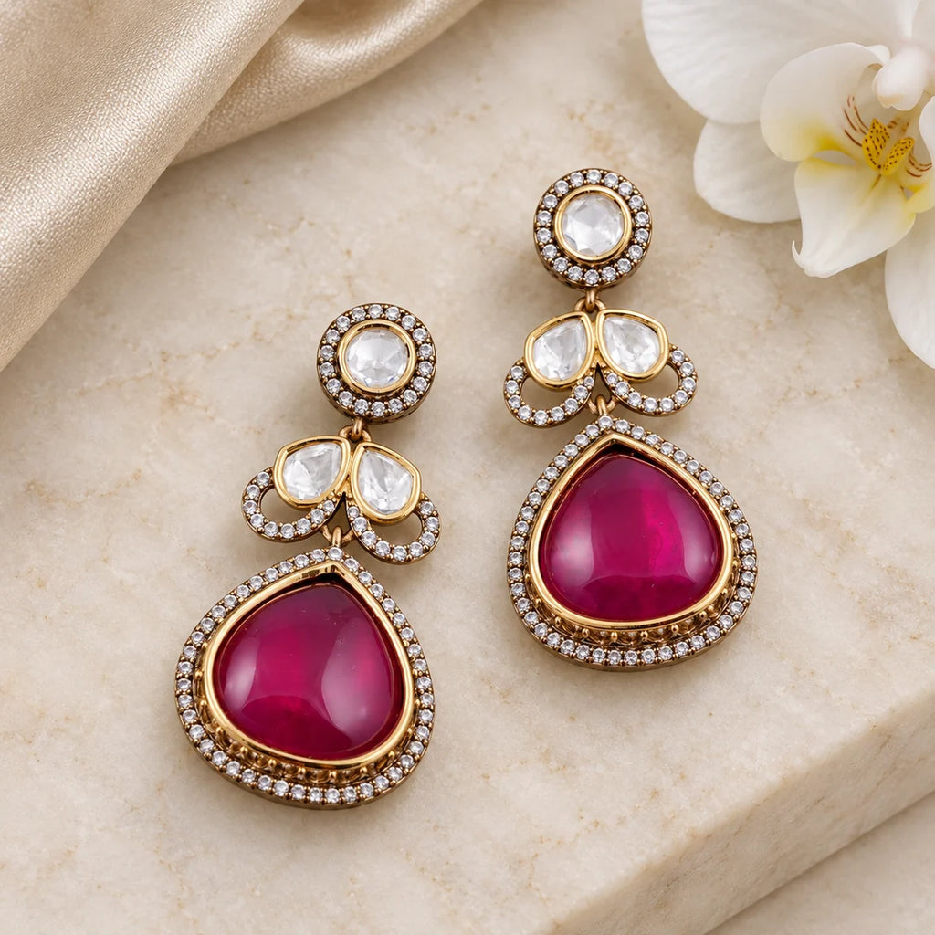 Pair of Prisha ruby-colour teardrop earrings on ivory stone beside champagne silk and a white orchid.
