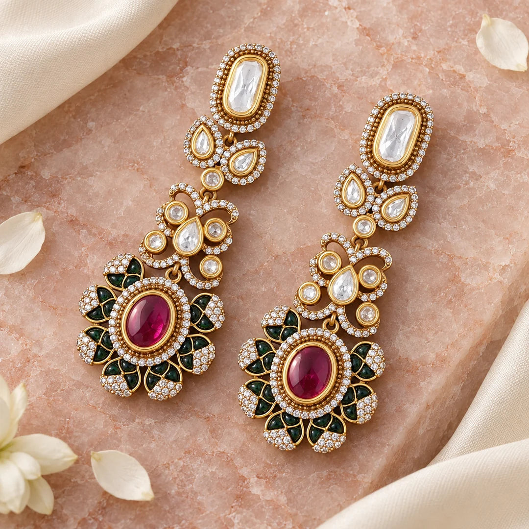 Pair of ruby-coloured and green long drop earrings lying flat on pale rose stone beside ivory silk and white petals.