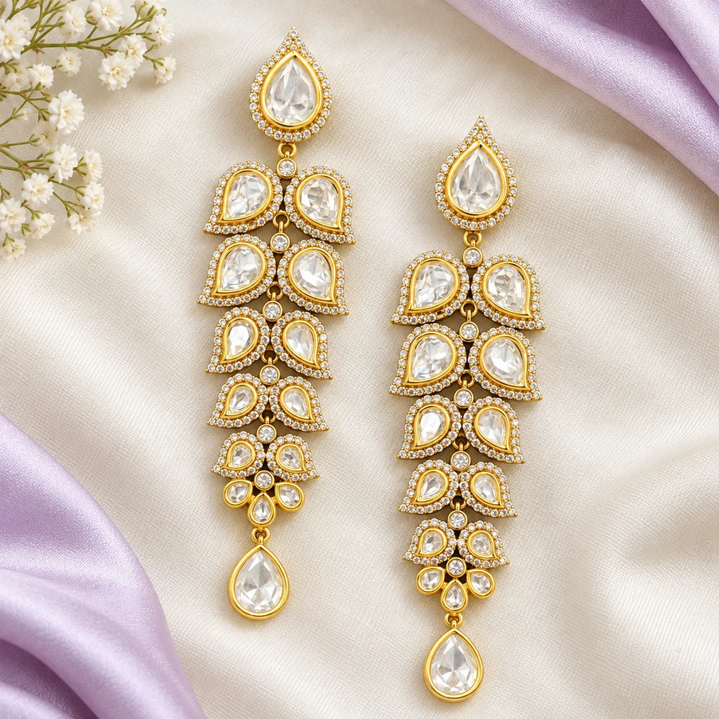 Pair of long clear-stone leaf earrings lying flat on ivory fabric framed by pale lavender silk and small white flowers.