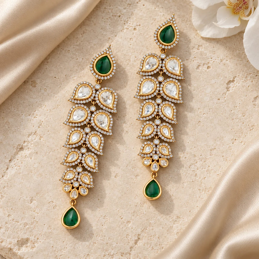 Pair of green and clear long leaf earrings lying flat on cream stone beside champagne silk and a white orchid.