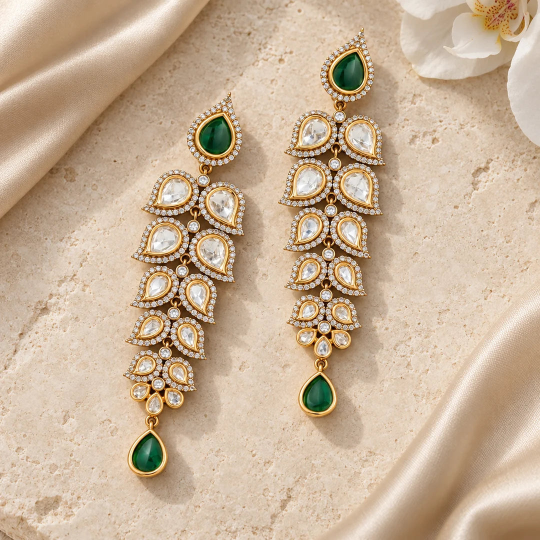 Pair of green and clear long leaf earrings lying flat on cream stone beside champagne silk and a white orchid.