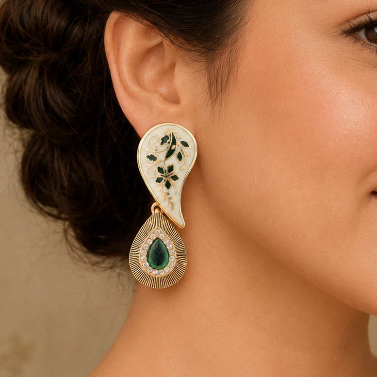 A close-up of a woman wearing a pair of gold and white teardrop-shaped earrings with green floral engravings and a green teardrop stone at the bottom.